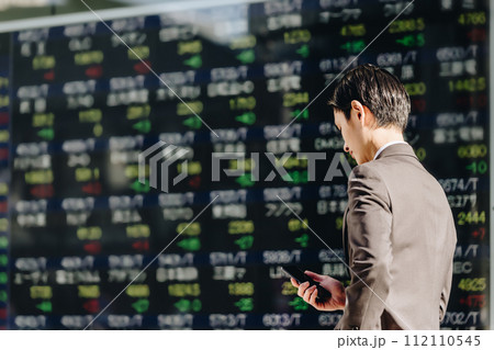 大量の株の銘柄が表示された株価ボードの前でスマートフォンを操作する30代の日本人の男性の後ろ姿 112110545