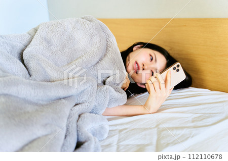 朝の寝起きに寝室でベッドに横たわりスマートフォンを見て寝不足や体調不良の為に寝坊した30代の女性 112110678