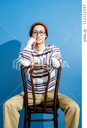 ブルーバックで椅子に座りながら考えているニット帽を被ったメガネの若い男性 112112547