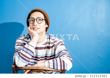 ブルーバックで椅子に座りながら考えているニット帽を被ったメガネの若い男性 112112561