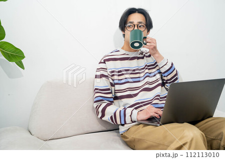 ソファでくつろぎながらコーヒーを飲みパソコンを操作する男性 ソファでくつろぎながらコーヒーを飲みパソコンを操作する男性 112113010