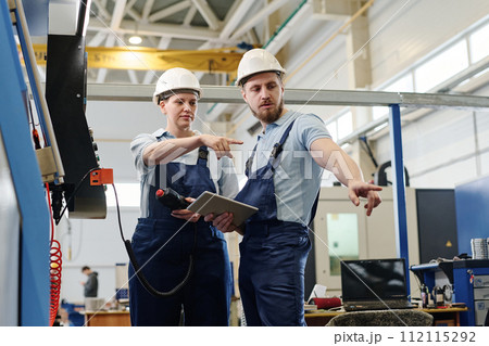 Low angle medium long shot of ethnically diverse young man and woman finding problem while working with CNC machine Low angle medium long shot of ethnically diverse young man and woman finding problem while working with CNC machine 112115292