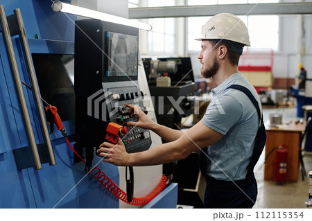 Side view of young bearded Caucasian man wearing hardhat operating modern CNC machine Side view of young bearded Caucasian man wearing hardhat operating modern CNC machine 112115354