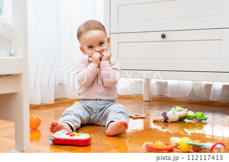 The baby hides his smile with his hands while sitting on the floor and playing with toys 112117413