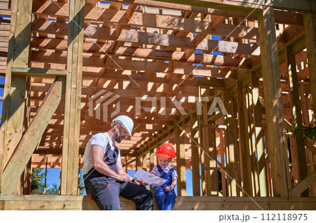 Father with toddler son building wooden frame house. Guys having fun on edge of balcony, examining construction plan, wearing helmets and overalls on sunny day. Carpentry and family concept. Father with toddler son building wooden frame house. Guys having fun on edge of balcony, examining construction plan, wearing helmets and overalls on sunny day. Carpentry and family concept. 112118975