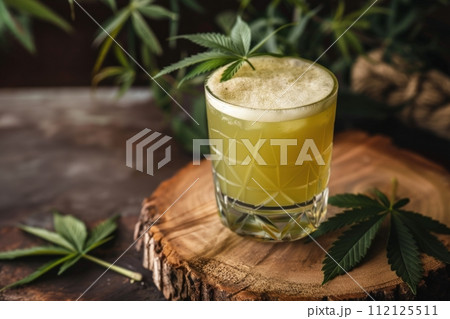 A refreshing cocktail in a glass topped with froth and a cannabis leaf on a rustic wooden slab A refreshing cocktail in a glass topped with froth and a cannabis leaf on a rustic wooden slab 112125511