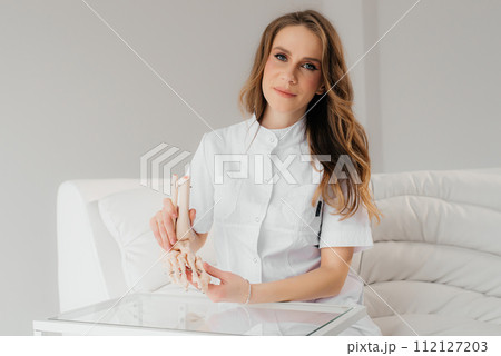 A doctor studies a womans leg structure while holding model of skeleton on a white table indoors. Models of skeletons help doctors study the structure of the human body A doctor studies a womans leg structure while holding model of skeleton on a white table indoors. Models of skeletons help doctors study the structure of the human body 112127203