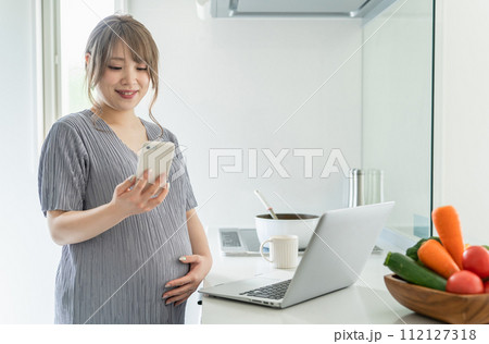 キッチンで料理をしながらパソコンとスマホを使う妊婦の女性 キッチンで料理をしながらパソコンとスマホを使う妊婦の女性 112127318