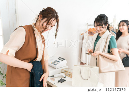 アパレルショップでレジでデニムを畳む女性ショップ店員とスマホを持って並ぶ女性客 112127376