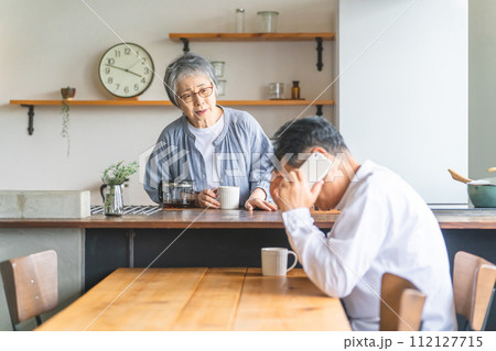自宅のダイニングで電話する夫を心配そうに見つめるキッチンにいるシニア女性・妻 112127715