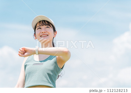 青空の下で腕時計を見ながら運動・トレーニング・ウォーキングする女性(歩数計・万歩計・心拍数) 112128213