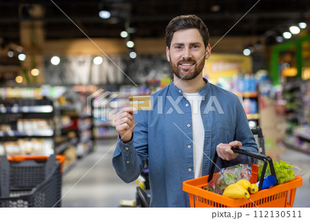 A young adult man holding a credit card and a basket full of fresh produce, ready for checkout at a modern supermarket. Emphasizing convenience and lifestyle. A young adult man holding a credit card and a basket full of fresh produce, ready for checkout at a modern supermarket. Emphasizing convenience and lifestyle. 112130511