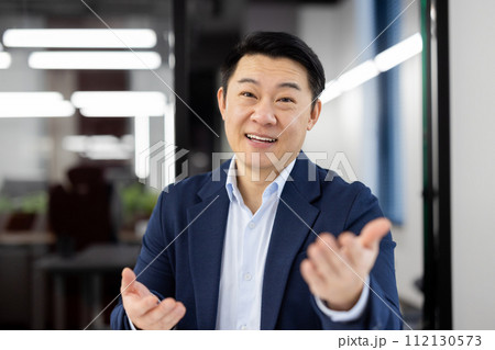Adult office administrator in business suit looking at camera with friendly smile and gesturing with hands. Hosting company receptionist welcoming partners of boss and asking to join in boardroom. Adult office administrator in business suit looking at camera with friendly smile and gesturing with hands. Hosting company receptionist welcoming partners of boss and asking to join in boardroom. 112130573