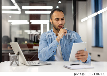 A professional male entrepreneur in a blue shirt is absorbed in reading financial reports on a digital tablet at his workspace, illustrating the concept of business analysis, digital workflow, and 112130714
