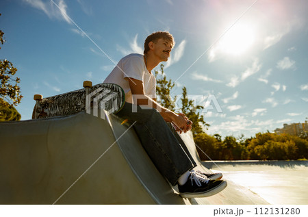 Low angle of young male skateboarder holding skateboard sitting on ramp in skate park  112131280