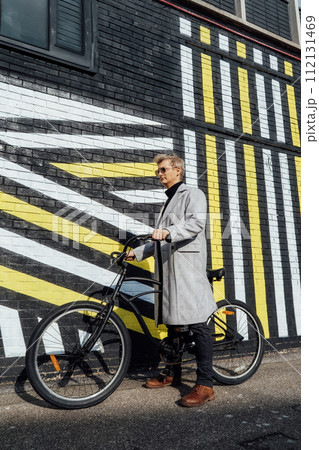 Stylish male in checked coat and sunglasses standing with retro bicycle by painted urban wall in sunny day. Neutral carbon footprint transportation. Green eco friendly mobility sustainable transport 112131469