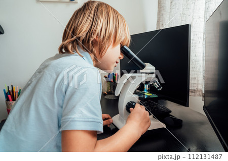 Caucasian boy using a microscope at home at his study place. Child curiosity, thirst for knowledge, home learning experience, home remote education concepts. Selective focus. Caucasian boy using a microscope at home at his study place. Child curiosity, thirst for knowledge, home learning experience, home remote education concepts. Selective focus. 112131487