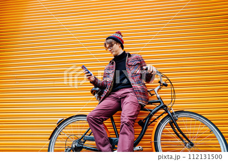 Young hipster man using mobile phone while sitting on cruiser bicycle near orange wall. Student using 5g network,rests after riding. Eco friendly transport for carbon footprint. Sustainable lifestyle 112131500