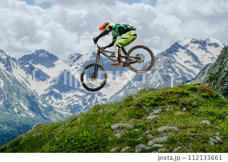 Mountain biker jumped, soaring mid-air against backdrop of snow-capped peaks and lush greenery. downhill competition 112133661