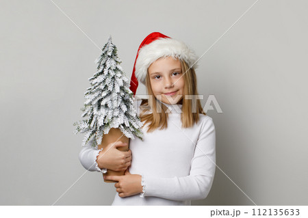 Young adorable child girl in Santa Claus hat posing with Christmas tree in her hands Young adorable child girl in Santa Claus hat posing with Christmas tree in her hands 112135633