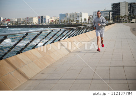 Full length portrait handsome young athletic man jogging, performing sprint run on running track in the coastline city 112137794