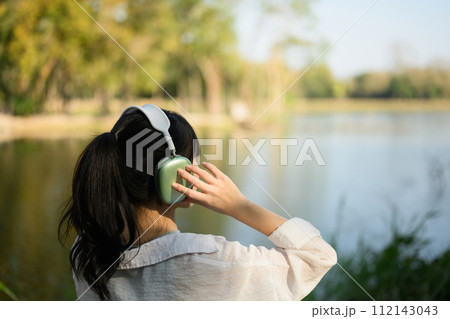 Back view of young woman listening to music in headphone and admiring lake in the summer park Back view of young woman listening to music in headphone and admiring lake in the summer park 112143043