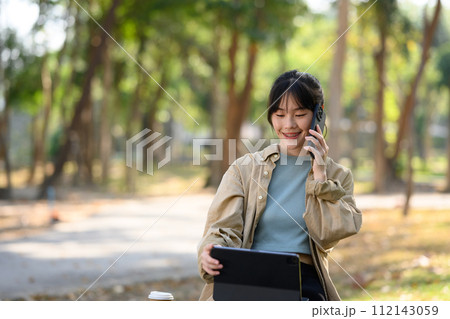 Smiling young woman freelancer sitting on park bench with digital tablet and talking on mobile phone Smiling young woman freelancer sitting on park bench with digital tablet and talking on mobile phone 112143059
