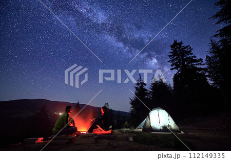 Night-time sojourn in mountains, beneath starry sky. Man and woman, intrepid wanderers, unwind by flickering flames of campfire, alongside their tent pitched near the serene forest, under Milky way. 112149335