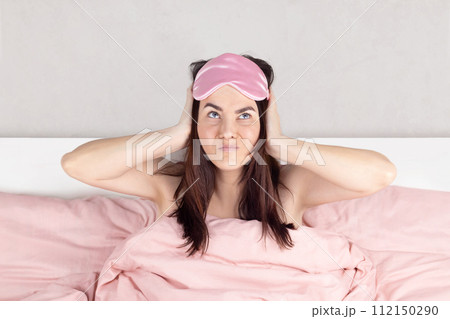 Woman cannot sleep, she covers her ears with her hands, the neighbors are noisy, making repairs. Pink sleep mask, pink bed linen. 112150290