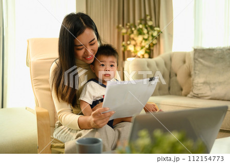 Smiling Asian mother working at home with laptop and documents sitting on armchair with cute little baby son 112154773