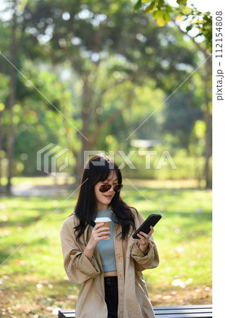 Portrait of young stylish woman in sunglasses drinking coffee and using mobile phone in the park Portrait of young stylish woman in sunglasses drinking coffee and using mobile phone in the park 112154808