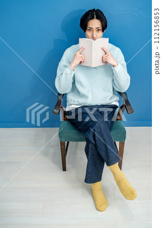 ブルーバックで椅子に座り本を読む若い男性 112156853