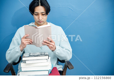 ブルーバックで椅子に座り本を読む若い男性 112156860
