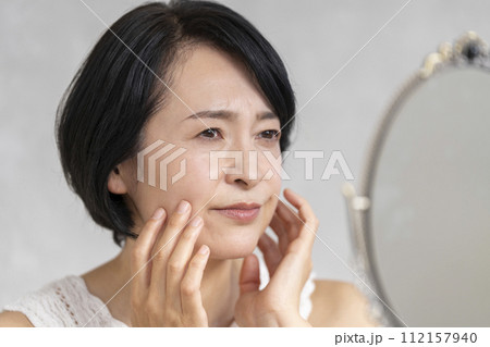鏡を見ながら顔に手をあてる50代前半の女性　悩み・肌トラブル　ビューティーイメージ 112157940