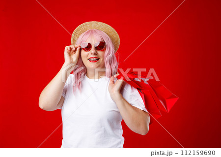 Portrait of an excited beautiful woman in hat and sunglasses holding red shopping bags isolated over red background. Black Friday, sale, buy day concept. Pink hair, studio shoot Portrait of an excited beautiful woman in hat and sunglasses holding red shopping bags isolated over red background. Black Friday, sale, buy day concept. Pink hair, studio shoot 112159690