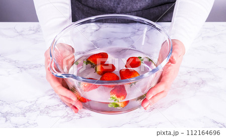 Ripe strawberries are submerged in water within a large glass mixing bowl, a step in washing the fruit to ensure cleanliness and longevity before storage or consumption. 112164696