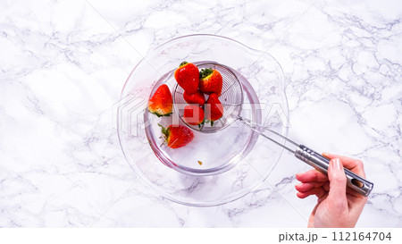 Flat lay. Ripe strawberries are submerged in water within a large glass mixing bowl, a step in washing the fruit to ensure cleanliness and longevity before storage or consumption. Flat lay. Ripe strawberries are submerged in water within a large glass mixing bowl, a step in washing the fruit to ensure cleanliness and longevity before storage or consumption. 112164704