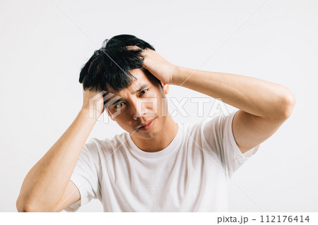Portrait of a young Asian man, looking sad and exhausted, holding his head in despair due to stress and a headache. Studio shot isolated on white, depicting suffering and depression. Portrait of a young Asian man, looking sad and exhausted, holding his head in despair due to stress and a headache. Studio shot isolated on white, depicting suffering and depression. 112176414