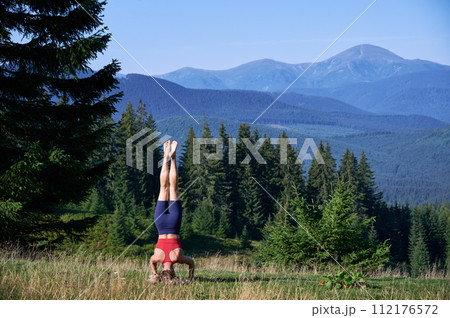 Woman doing morning exersices in mountains. Slim lady training outdoors, wearing sportswear in summer. Flexible woman standing on head on grass, doing yoga. Concept of harmony with nature. 112176572