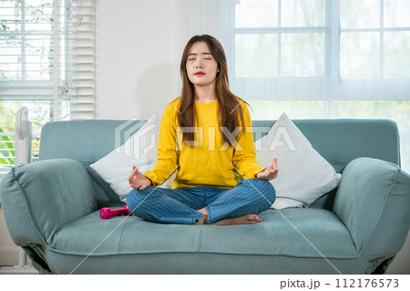 Young woman practise yoga and meditation in lotus position, mindfulness lifestyle, Asian female sits on sofa at home and chilling with couch lotus pose eyes closed, happy relaxation leisure people 112176573