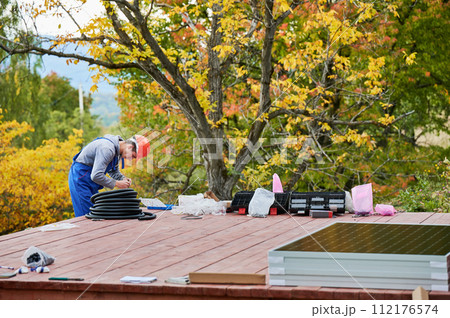 Man worker prepearing for mounting photovoltaic solar modules on roof of house. Electrician in helmet installing solar panel system outdoors. Concept of alternative and renewable energy. 112176574