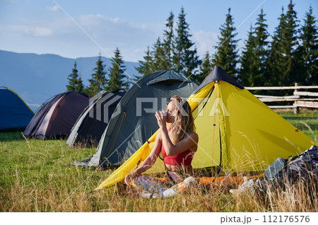 Woman hiker camping outdoors. Tourist sitting in campsite, admiring landscape and drinking tea. Sporty, young female having break, enjoying in mountains. Concept of beauty of nature. 112176576