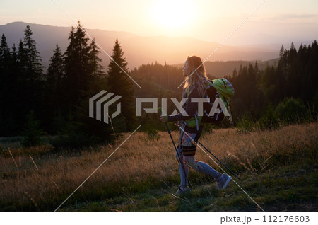 Woman hiker hiking outdoors at sunset. Sporty, positive woman traveling in mountains. Young, sporty female with backpack using trekking sticks, walking. Concept of harmony with nature. Woman hiker hiking outdoors at sunset. Sporty, positive woman traveling in mountains. Young, sporty female with backpack using trekking sticks, walking. Concept of harmony with nature. 112176603