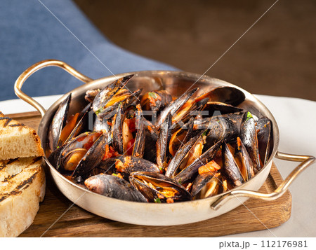 Delicious black mussels in red tomato sauce. Mediterranean black mussels in metal pan on wooden cutting board with grilled bread on restaurant table background Delicious black mussels in red tomato sauce. Mediterranean black mussels in metal pan on wooden cutting board with grilled bread on restaurant table background 112176981