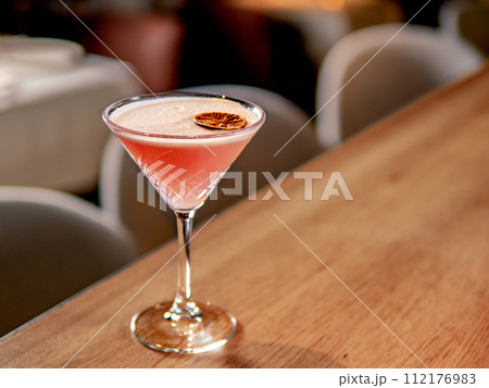 Red cocktail in martini glass on bar counter in restaurant interior. Gin alcohol cocktail decorated egg white and dry lichee fruit slice. Pink cold alcoholic cocktail in bar or nightclub background 112176983
