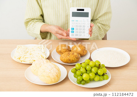 糖質を多く含む食品(炭水化物)と電卓を持つミドル女性 糖質を多く含む食品(炭水化物)と電卓を持つミドル女性 112184472