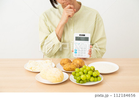 糖質を多く含む食品(炭水化物)と電卓を持って悩むミドル女性 糖質を多く含む食品(炭水化物)と電卓を持って悩むミドル女性 112184656