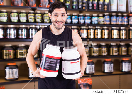 Young bodybuilder showing power and holding pot of sport nutrition products in shop 112187555