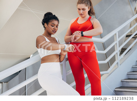 Two plus-size women in fitness attire are checking pulse during a workout. Young females in sports clothes are standing on stairs outdoors looking at smartwatches. Two plus-size women in fitness attire are checking pulse during a workout. Young females in sports clothes are standing on stairs outdoors looking at smartwatches. 112189238
