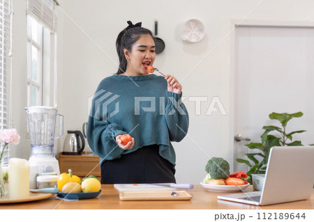 Woman eats a tomato by the laptop on the table by the window in the wood house Woman eats a tomato by the laptop on the table by the window in the wood house 112189644